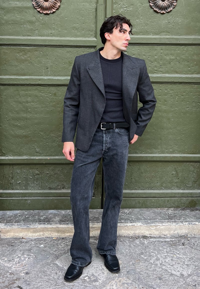 Blazer gris sur un haut noir, associé à un jean évasé en denim foncé et des chaussures noires. Tissu texturé et design structuré, debout devant un fond vert.
