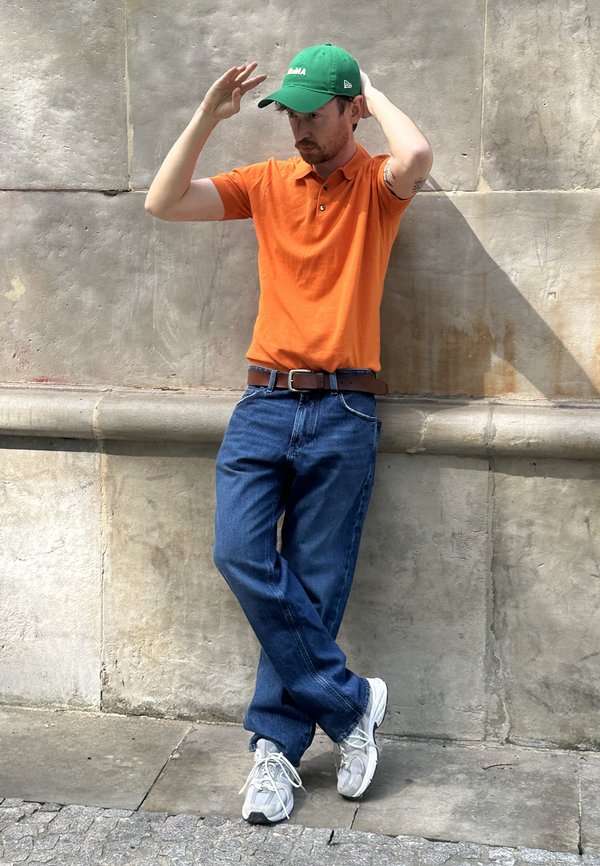 Uomo con berretto verde, polo arancione, jeans blu e scarpe da ginnastica bianche appoggiato a un muro di pietra, che si sistema il berretto con entrambe le mani.