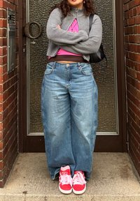 Cardigan gris à fermeture éclair, haut rose, jeans bleus oversize, et baskets rouges avec des lacets roses. Debout devant une porte en verre texturé.