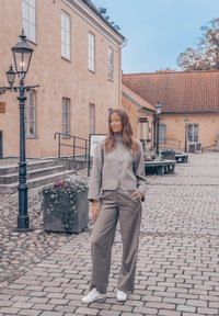 Γκρι ζακέτα με φερμουάρ και ταιριαστά φαρδιά παντελόνια, συνδυασμένα με λευκά sneakers; μπροστά σε έναν λιθόστρωτο δρόμο και ιστορικά κτίρια.