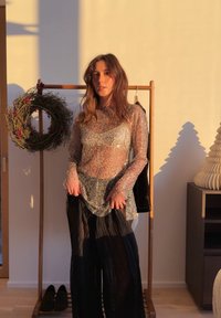 Femme en haut transparent à sequins et pantalon large noir, debout à l'intérieur près d'un portant en bois avec une couronne et une décoration éclairée par des ombres.