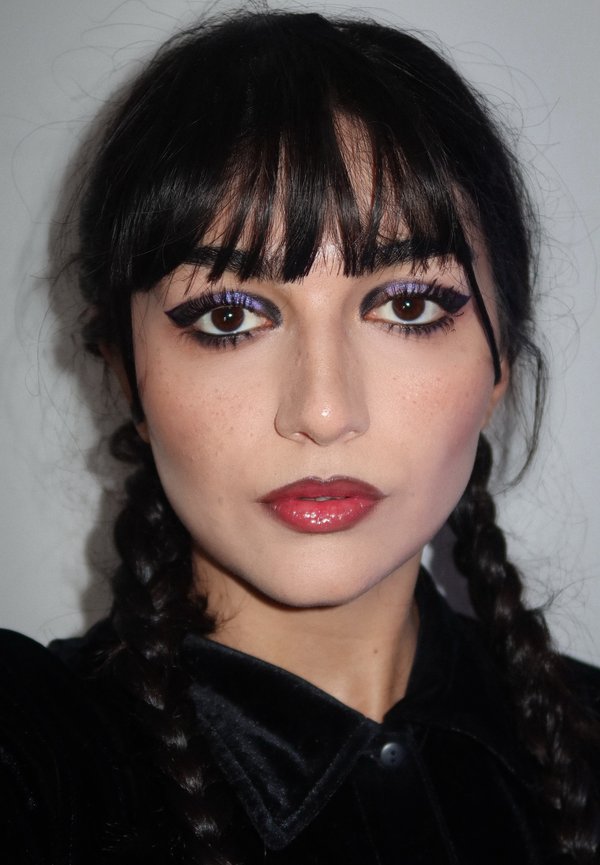 Portrait d'une personne aux cheveux noirs tressés en deux nattes, portant une chemise noire en velours avec col, et affichant un maquillage des yeux audacieux avec des accents violets.