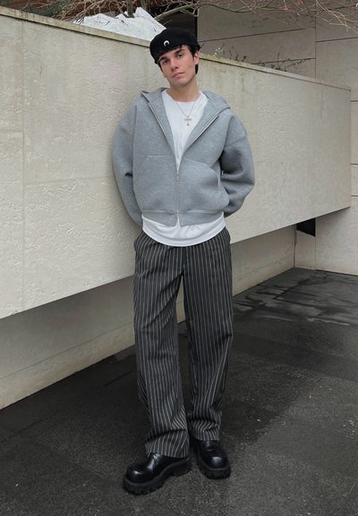 Sudadera gris con cremallera sobre una camiseta blanca, combinada con pantalones a rayas blancos y negros y zapatos negros gruesos. Texturas suaves, ajuste casual.