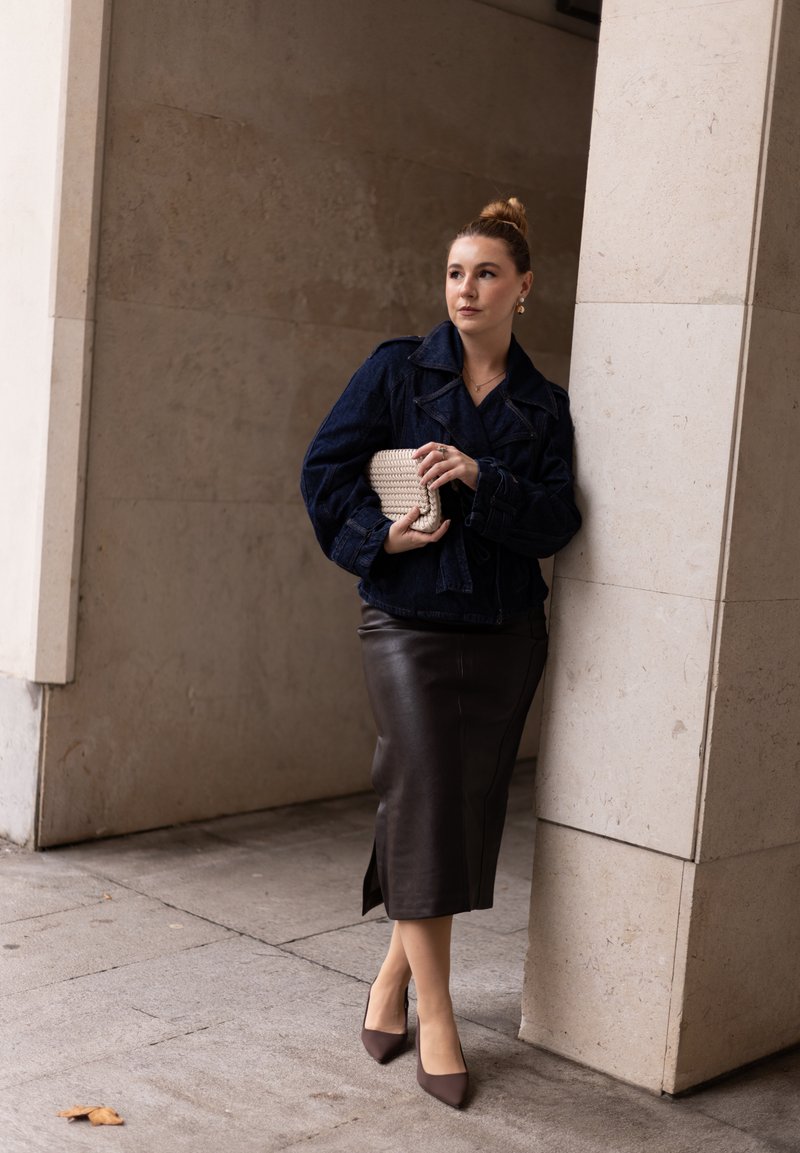 Veste en denim, jupe midi en cuir marron foncé avec une fente sur le côté, pochette tissée beige et escarpins à bout pointu marron. Textures lisses, coupe ajustée.