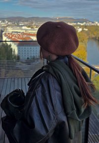 Smešasti smešak, smeđe pletivo beretka, crna kožna jakna, zeleni šal, drži crnu torbu. Urbani pejzaž sa zgradama i reku u pozadini.