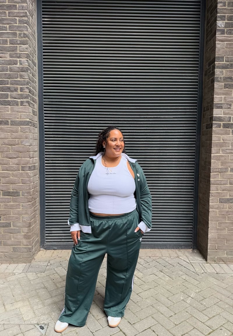 Mulher sorridente com cabelo encaracolado está em frente a uma persiana de metal preta, vestindo um fato de treino verde, um top curto branco e ténis, com as mãos nos bolsos.