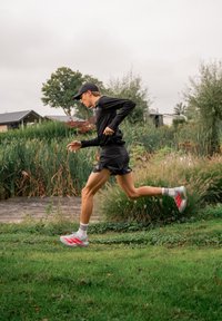 Runner in abbigliamento sportivo nero e cappellino in corsa su un sentiero erboso vicino a canne alte e piccole case sotto un cielo nuvoloso.