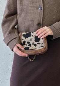 Καφέ τσάντα με καρό σχέδιο, με καμβά μπροστά που φέρει μαύρο και μπεζ σχέδιο γελάδας, χρυσή αλυσίδα και λεπτομέρεια λογότυπου.