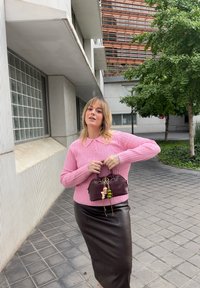 Różowy prążkowany sweter z kołnierzykiem, czarna skórzana spódnica ołówkowa i mała burgundowa torebka z kolorowymi zawieszkami. Teksturowane otoczenie na zewnątrz.