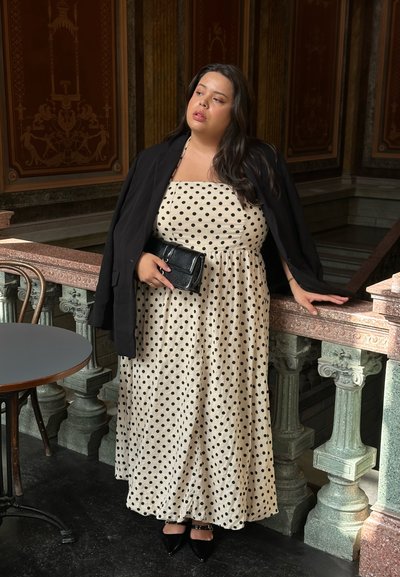 Vestido crema con lunares negros; presenta un diseño sin tirantes, una blazer negra y un bolso negro brillante. Los tacones puntiagudos completan el look.