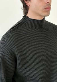 Mann iført en mørk ribbestrikket genser med høy hals og en liten svart klemme nær kragen mot en ensfarget lys bakgrunn.