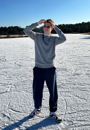 Suéter gris, pantalones azul marino y zapatillas negro y rojo. De pie sobre la nieve, usando gafas de sol redondas, con un cielo azul despejado de fondo.