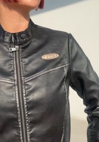 Czarna skórzana kurtka motocyklowa z wysokim kołnierzem, zamkiem błyskawicznym i małym haftowanym logo na lewej piersi. Gładka tekstura i dopasowany krój.