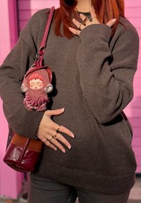 Γυναίκα με πουλόβερ γκρι πλεκτό και κόκκινα μαλλιά κρατάει μπορντό τσάντα με μπρελόκ λούτρινης κούκλας, φοράει πολλά δαχτυλίδια, στέκεται μπροστά σε ροζ τοίχο.