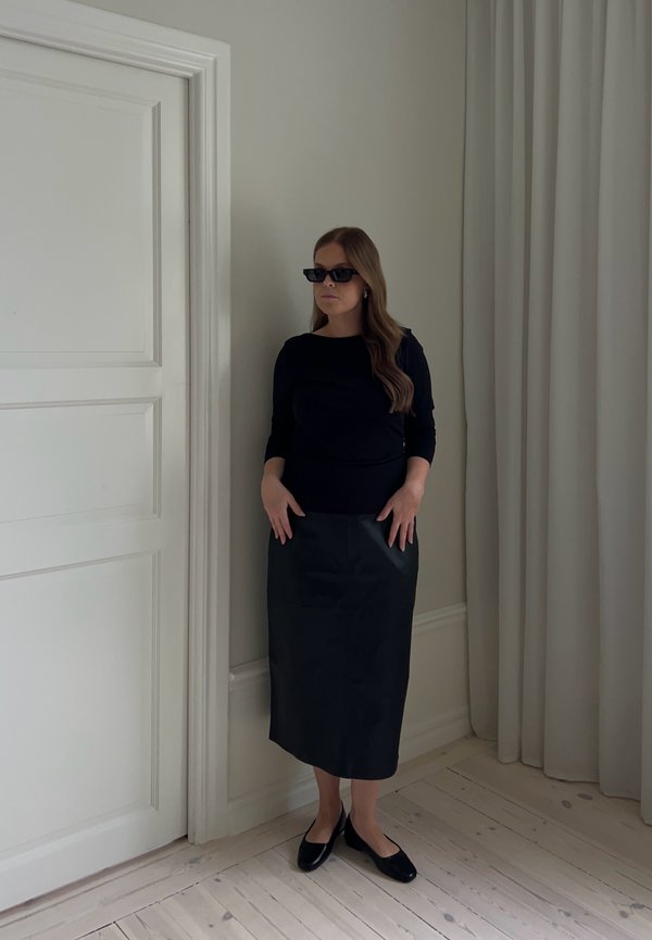 Donna con capelli lunghi che indossa occhiali da sole neri, top nero, gonna midi nera e scarpe nere, in piedi all'interno vicino a una porta bianca e tende.
