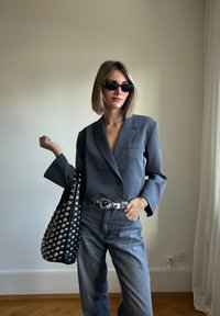 Šedý pruhovaný blazer, džíny s vysokým pasem tmavé barvy, černá kabelka s hřebíky a černé sluneční brýle. Hladká textura, přizpůsobený střih, strukturovaný design.