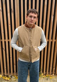 Béžová prošívaná vesta s hnedým golierom, sivé tričko s dlhým rukávom a modré nohavice s vzorom. Dve predné vrecká a zapínanie na zips. Pozadie z drevených lamiel.