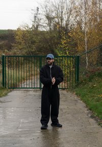 Čierna oversize bunda na zips, rovnaké široké nohavice a čierne topánky. Modrá čiapka a okuliare dopĺňajú outfit na pozadí zelených brán.