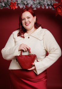 Sac à main en cuir rouge de forme triangulaire avec de petites poignées, présentant une finition texturée. Accompagné d'un pull en maille crème avec un col orné de bijoux.