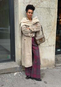 Manteau long en suédine beige, chemise blanche, pantalons larges à carreaux bordeaux et verts, sac en cuir marron, chaussures sombres, debout contre un mur en pierre.
