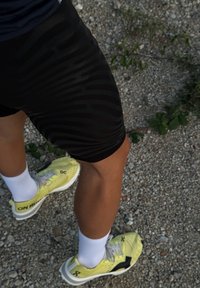 Svarta sportshorts med ett vågmönster, i kombination med gula löparskor och vita strumpor. Underlaget är grus med liten grönska.
