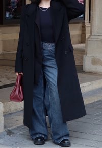 Zwarte wollen overjas, marineblauw nauwsluitend topje, wijde blauwe jeans en stevige zwarte laarzen. Houdt een kleine rode handtas vast. Soepele texturen, op maat gemaakt ontwerp.