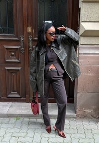 Överdimensionerad mörkgrön läderjacka, svart topp, ribbade bruna byxor, röd handväska och vinröda klackar; texturerad bakgrund med en träddörr.