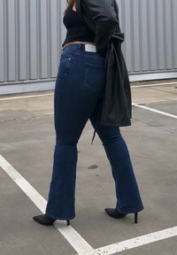 Jeans de ganga escuros, tipo flare, com cintura alta e bolso traseiro, combinados com sapatos de salto agudo pretos e um longo casaco preto, num ambiente ao ar livre.