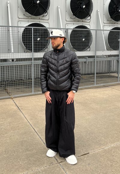 Hombre con chaqueta acolchada negra, pantalones anchos negros, zapatillas blancas y gorra blanca, de pie sobre cemento cerca de ventiladores industriales detrás de una reja metálica.