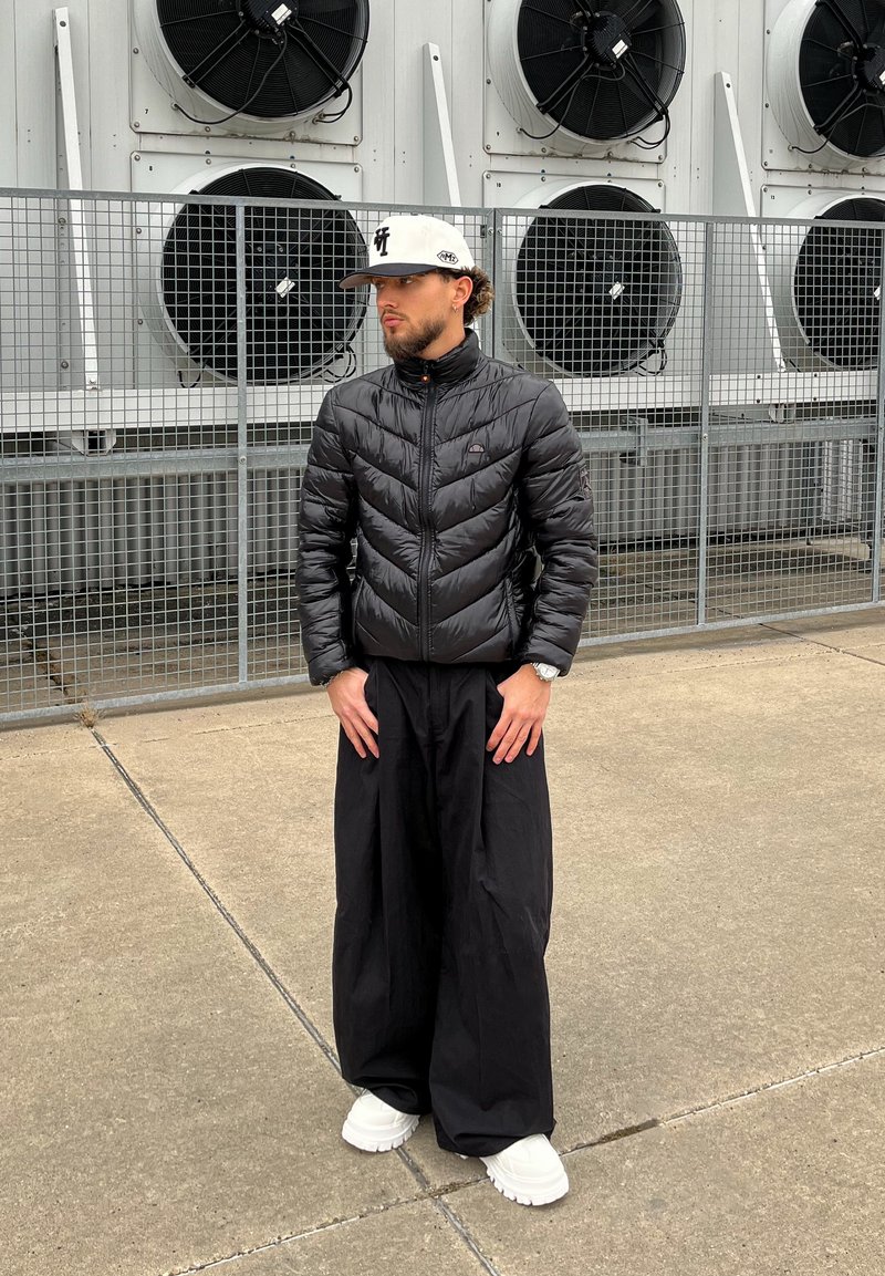 Homme en doudoune noire, pantalons larges noirs, baskets blanches et casquette blanche, debout sur du béton près de ventilateurs industriels derrière une clôture métallique.