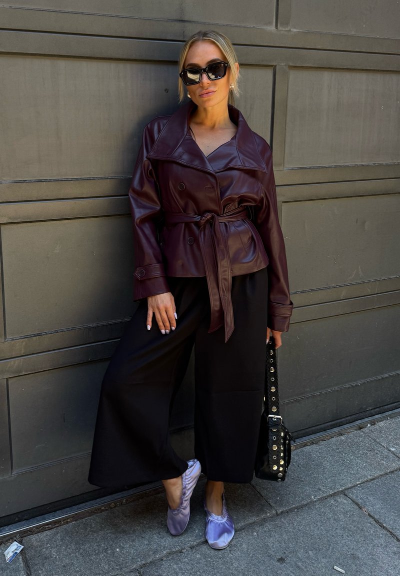 Femme portant une veste en cuir violet foncé et un pantalon large noir, lunettes de soleil, tenant un sac noir clouté, debout contre un mur gris.