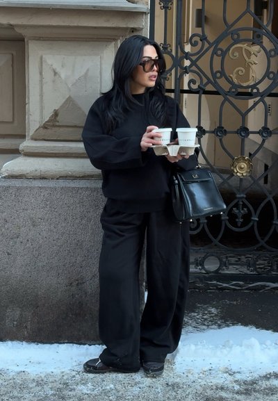 Mujer vestida de negro con gafas de sol sostiene cuatro tazas de café en una bandeja de cartón, de pie en una acera nevada cerca de un edificio de piedra y una puerta de hierro.