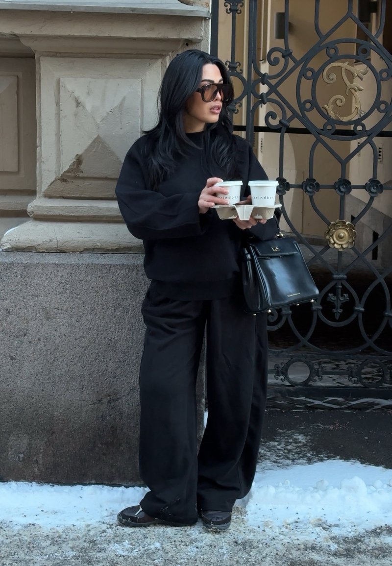 Femme vêtue entièrement de noir avec des lunettes de soleil tenant quatre tasses de café dans un plateau en carton, debout sur un trottoir enneigé près d'un bâtiment en pierre et d'une grille en fer.