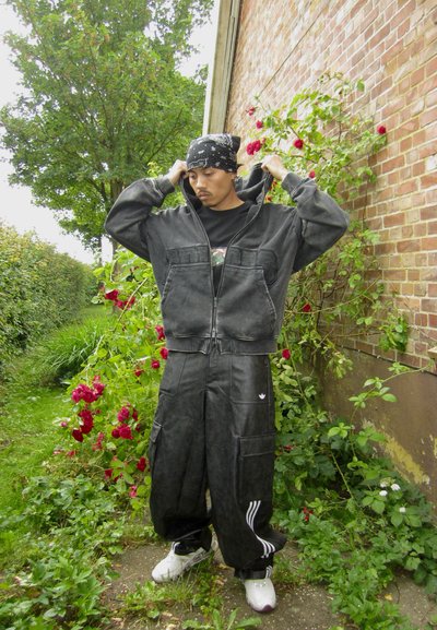 Chaqueta de mezclilla negra con capucha, cierre de cremallera y bolsillos frontales, combinada con pantalones cargo negros holgados que tienen rayas blancas y múltiples bolsillos.