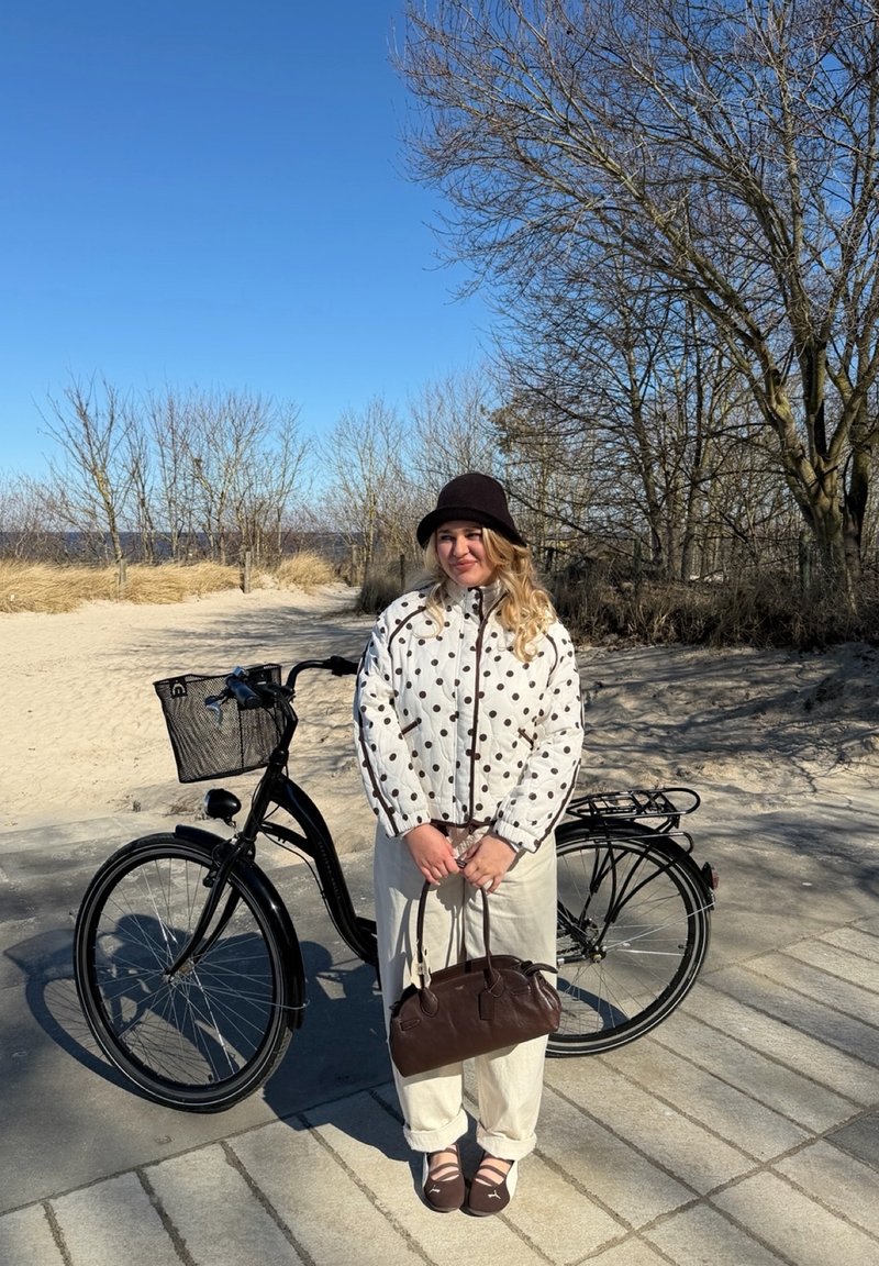 Junge Frau in einem gepunkteten Jackett und braunem Hut steht neben einem schwarzen Fahrrad und hält eine braune Handtasche in einer sonnigen Außenumgebung mit kahlen Bäumen.