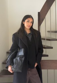 Egy személy által tartott fekete bőr válltáska, hosszú fekete kabáttal, amely egy sötét pulóver és egy szürke, galléros ing rétege fölött van. A háttérben fából készült lépcső.