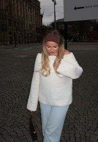 Cremefarbenes Off-Shoulder-Pullover mit langen Ärmeln, kombiniert mit hellblauen Hosen. Ein braunes Haarband und eine braune Handtasche runden das Outfit ab. Pflasterstein-Hintergrund.