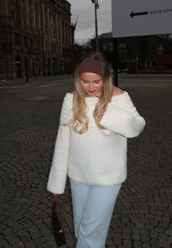 Cremefarbenes Off-Shoulder-Pullover mit langen Ärmeln, kombiniert mit hellblauen Hosen. Ein braunes Haarband und eine braune Handtasche runden das Outfit ab. Pflasterstein-Hintergrund.