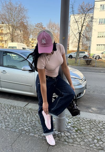 Camiseta ajustada de color rosa claro, vaqueros de pierna ancha azules oscuros, zapatillas rosas y una gorra de béisbol rosa con un logo blanco; bolso negro. Calle de adoquines.
