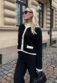 Ξανθιά γυναίκα που φοράει μαύρα γυαλιά ηλίου, μαύρο και λευκό ζακέτα με κουμπιά, μαύρο παντελόνι, κολιέ με φύλλα χρυσού, κρατώντας μαύρη τσάντα σε λιθόστρωτο δρόμο.