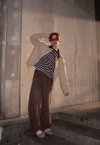 Bēša jaka, melnbalts svītrains krekls, brūnas plaša piegriezuma bikses, pelēkas zamšādas kurpes, apaļas saulesbrilles, stāv uz betona pakāpieniem.