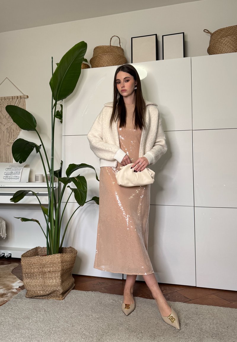 Frau in beigem Midikleid mit Pailletten und weißer flauschiger Jacke hält eine cremefarbene Clutch, steht neben einer großen grünen Pflanze und weißen Schränken in einem modernen Raum.