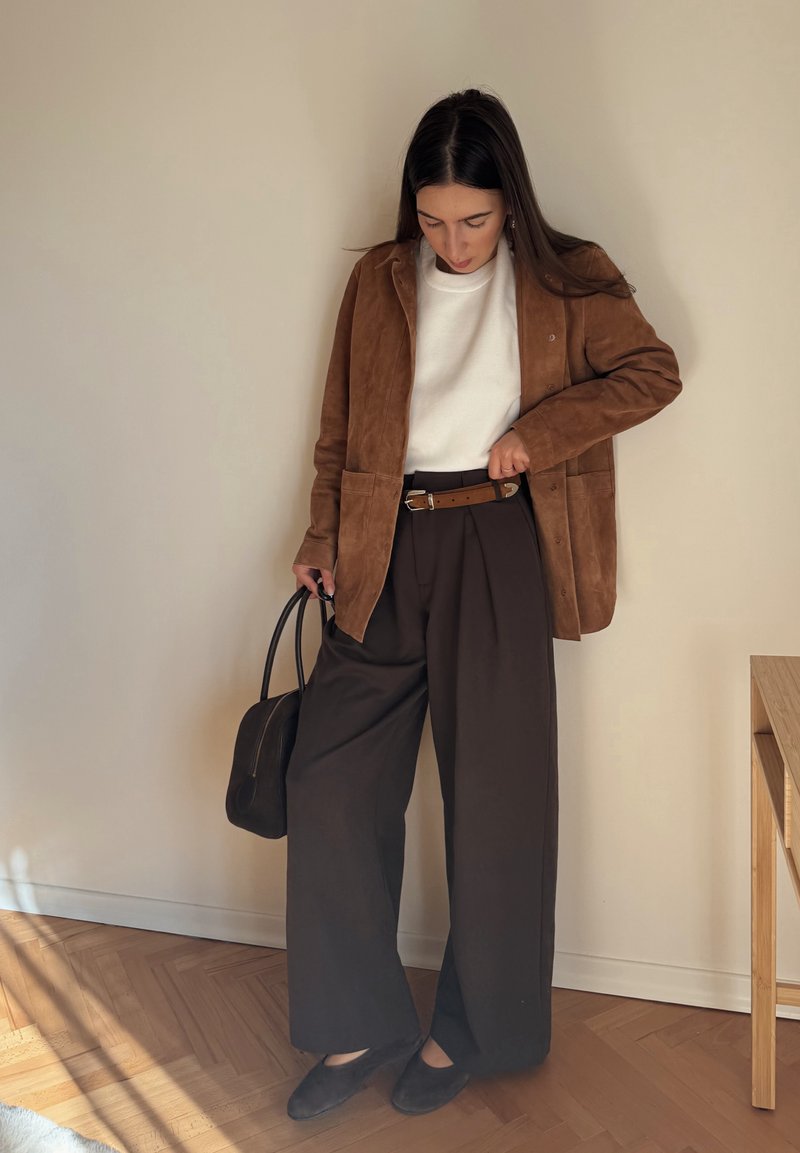Brown suede jacket over a white sweater, brown pleated trousers, brown belt, and black bag. Smooth textures; light and shadow play on surfaces.