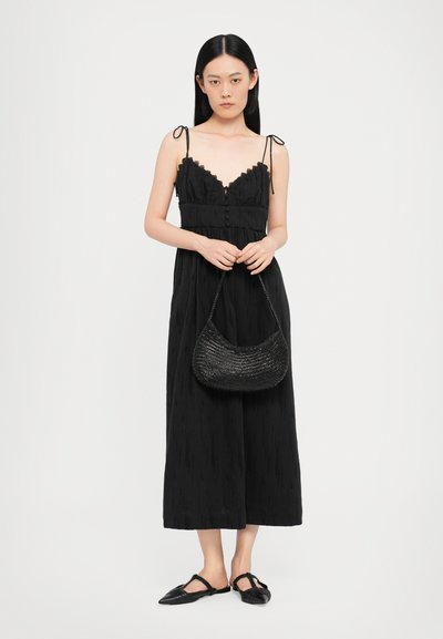 Vestido maxi negro sin mangas con ribete de encaje, un frente abotonado y tirantes ajustables. Lleva un bolso de mano tejido negro y zapatos planos.