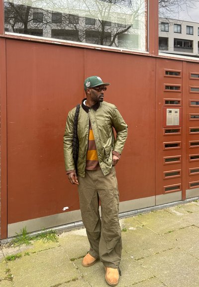 Chaqueta bomber verde sobre un suéter de rayas, pantalones cargo beige y botas marrones. De pie contra una pared marrón con un intercomunicador metálico.