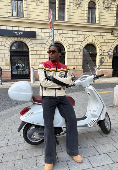 Chaqueta de motocicleta de cuero blanca y roja con acentos negros, jeans oscuros de pierna acampanada y botas marrones, de pie junto a un scooter blanco contra un edificio de piedra.