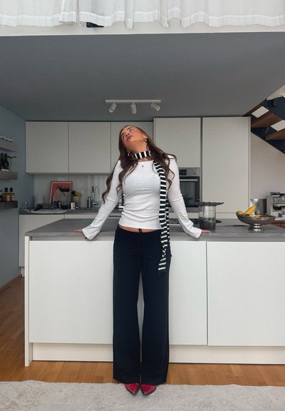Top blanco de manga larga con mangas acampanadas, pantalones negros de pierna ancha y bufanda de rayas blancas y negras. Tacones rojos de punta afilada. Fondo de cocina moderna.