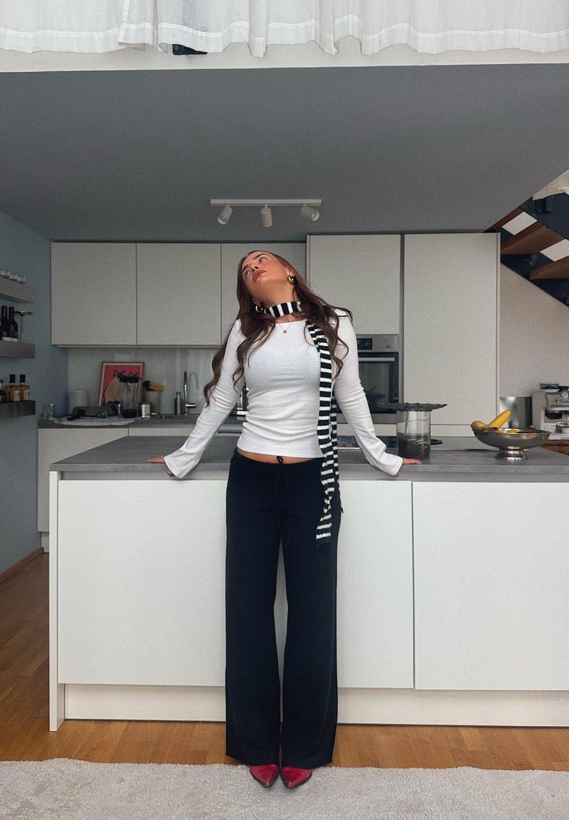 Haut blanc à manches longues avec manches évasées, pantalon large noir, et écharpe rayée noir et blanc. Escarpins rouges à bouts pointus. Décor de cuisine moderne.