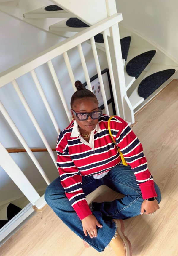 Červeno-námořnický pruhovaný polo tričko s dlouhým rukávem, modré džíny a béžové boty. Doplňky zahrnují žlutou tašku a vrstvené zlaté náhrdelníky.