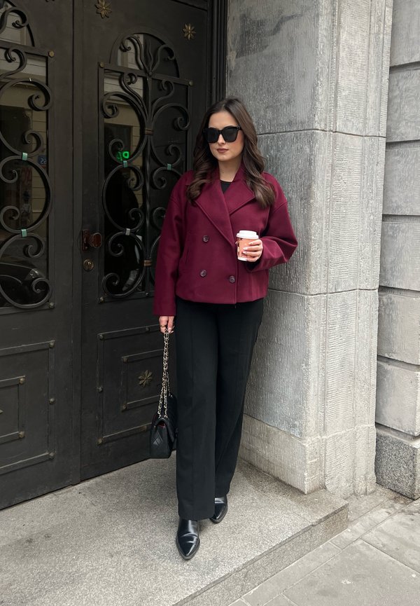 Cappotto bordeaux doppiopetto, pantaloni neri aderenti, scarpe nere a punta, con in mano una tazza di caffè, in piedi accanto a porte metalliche decorative.